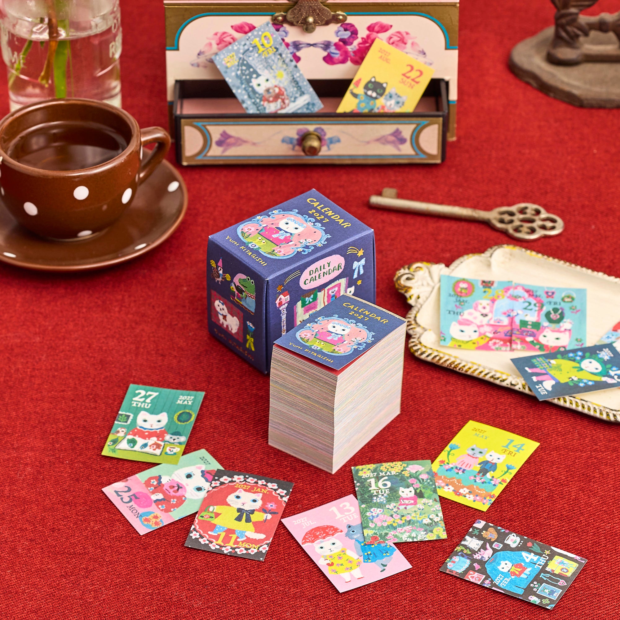 Calendario mini Yumi Kitagishi 2027 con caja y hojas ilustradas sobre una mesa roja